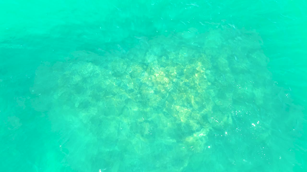 Sunlit rocks are visible below turquoise seawater at Marquis Los Cabos, with the brightest reflections clustered near the lower part of the frame and no visible people.