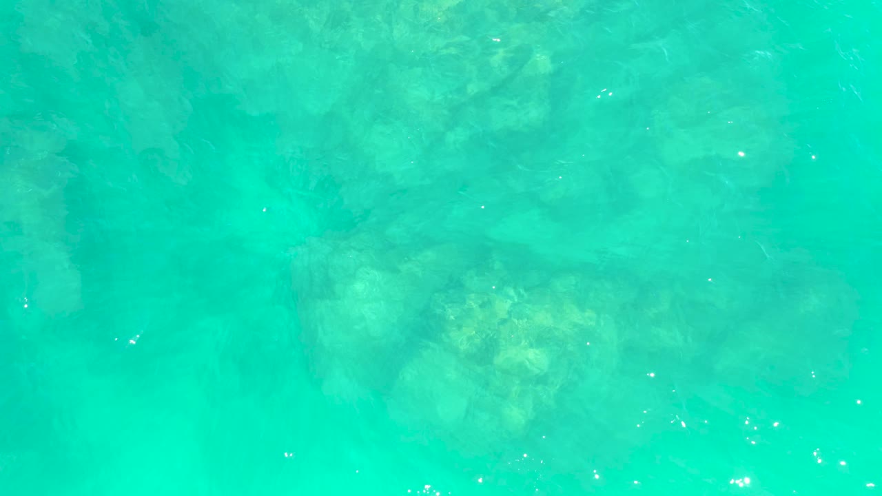 Clear turquoise water at Marquis Los Cabos with a pale rock cluster under the center-left area and small surface glints, no visible people.