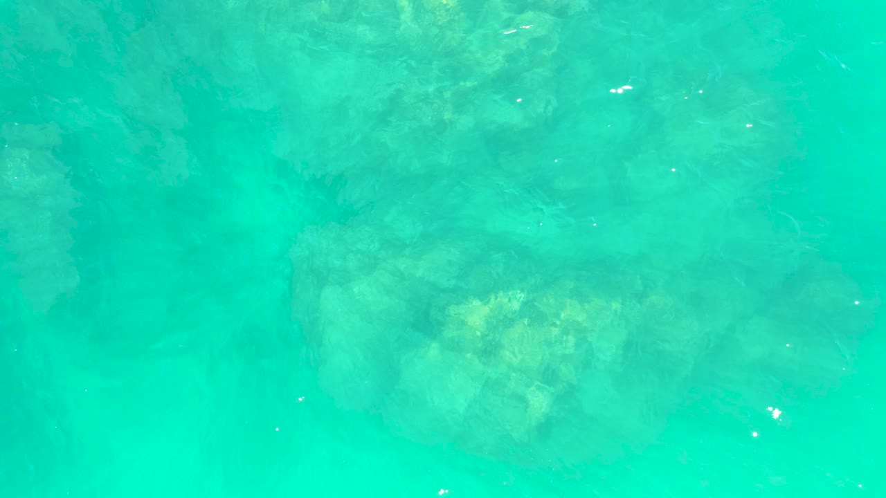 Blue-green shallow water at Marquis Los Cabos with muted rocky forms below the surface and scattered bright reflections, no visible people.