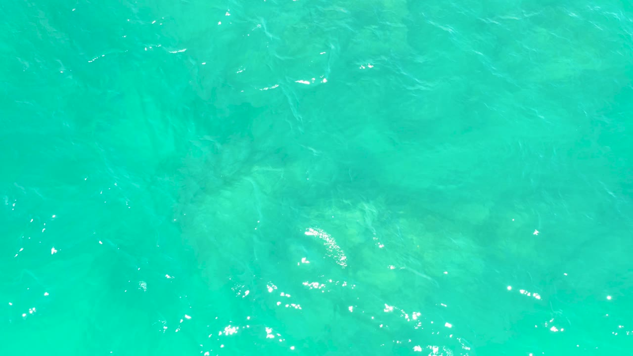 Turquoise ocean surface at Marquis Los Cabos carries brighter sun flecks over muted rock shapes below, with no visible people.
