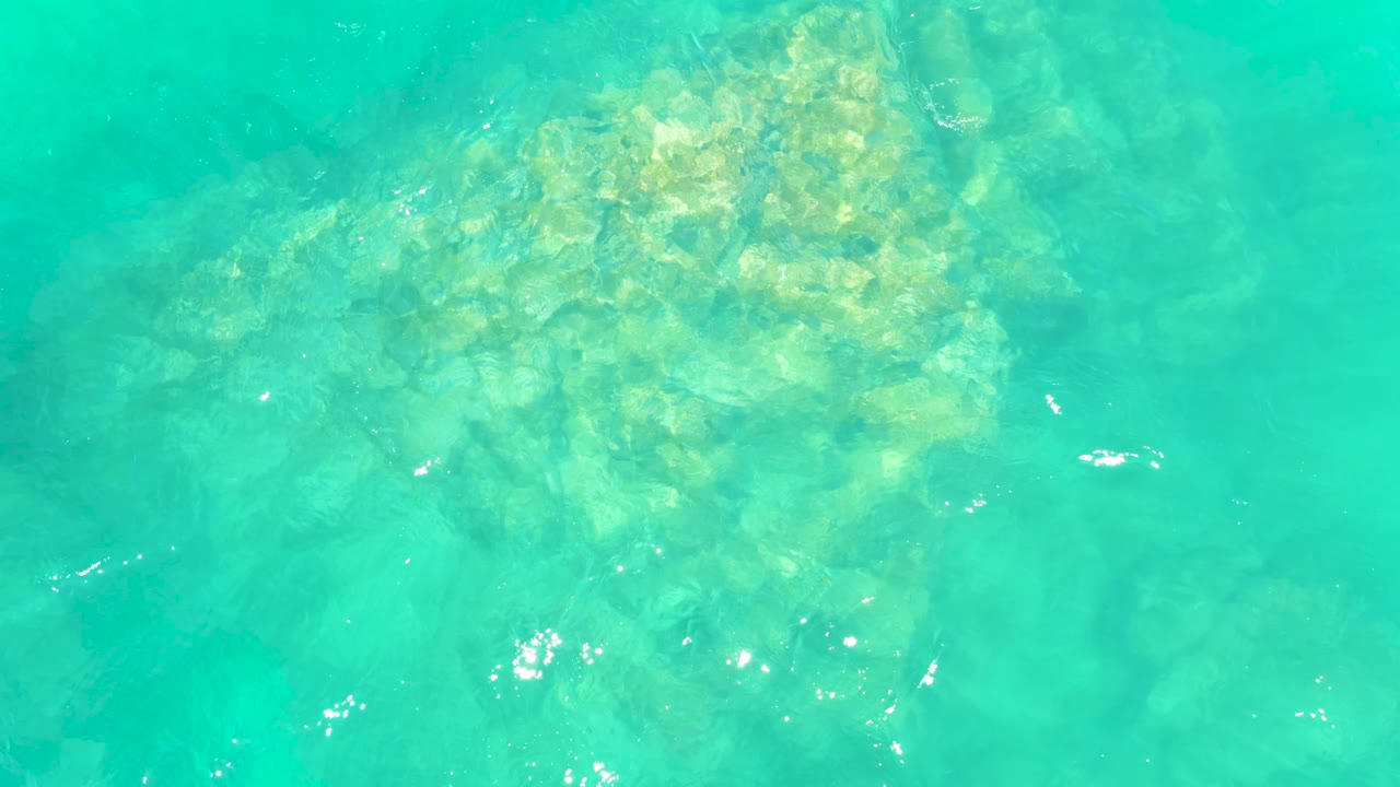 A pale rock formation sits under bright turquoise water at Marquis Los Cabos, with small white reflections on the surface and no visible people.