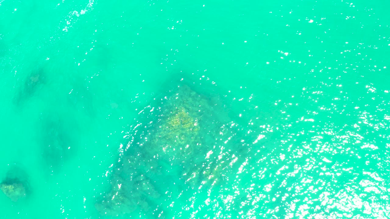 Overhead view of turquoise water at Marquis Los Cabos with submerged rock or coral shapes under ripples, bright surface highlights, and no visible people.