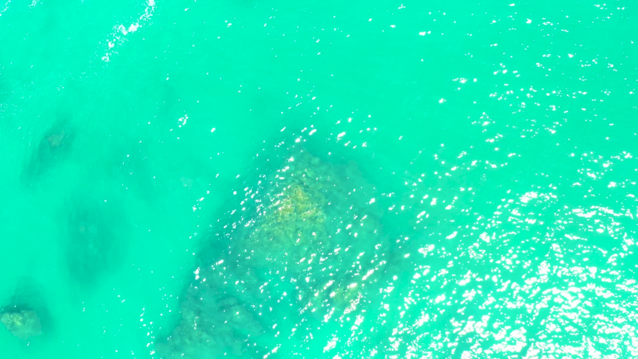 Overhead Marquis Los Cabos water detail with a larger submerged rock or coral patch near center-left, green-blue water, surface shimmer, and no visible people.