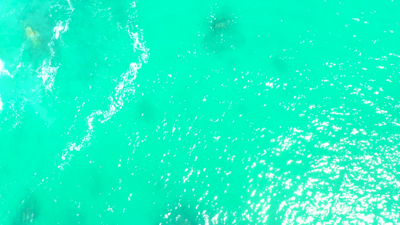 Top-down view of turquoise water at Marquis Los Cabos with white foam along the left edge, faint underwater rocks, bright sun reflection, and no visible people.