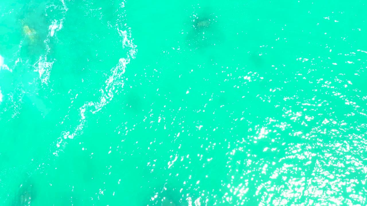 Aerial view of clear turquoise water at Marquis Los Cabos with a curved foam streak near the upper left, underwater rock shadows, strong surface sparkle, and no visible people.