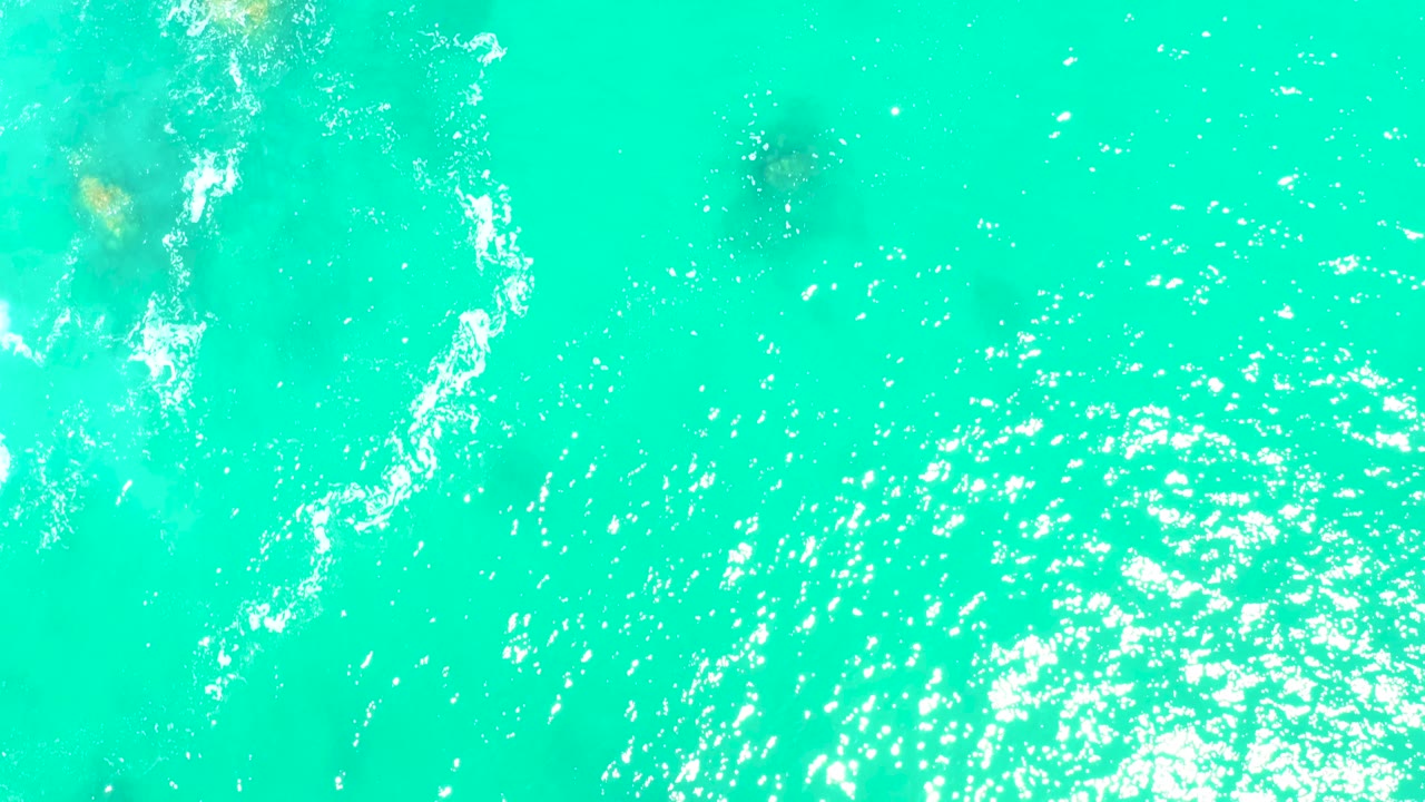 Top-down Marquis Los Cabos water detail with scattered white foam near the top, green-brown rocks visible below the turquoise surface, sun flecks, and no visible people.