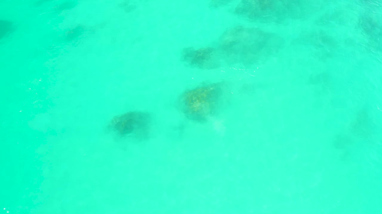 A high view of blue-green water at Marquis Los Cabos reveals muted submerged rocks across the frame beneath gentle ripple patterns, no visible people.