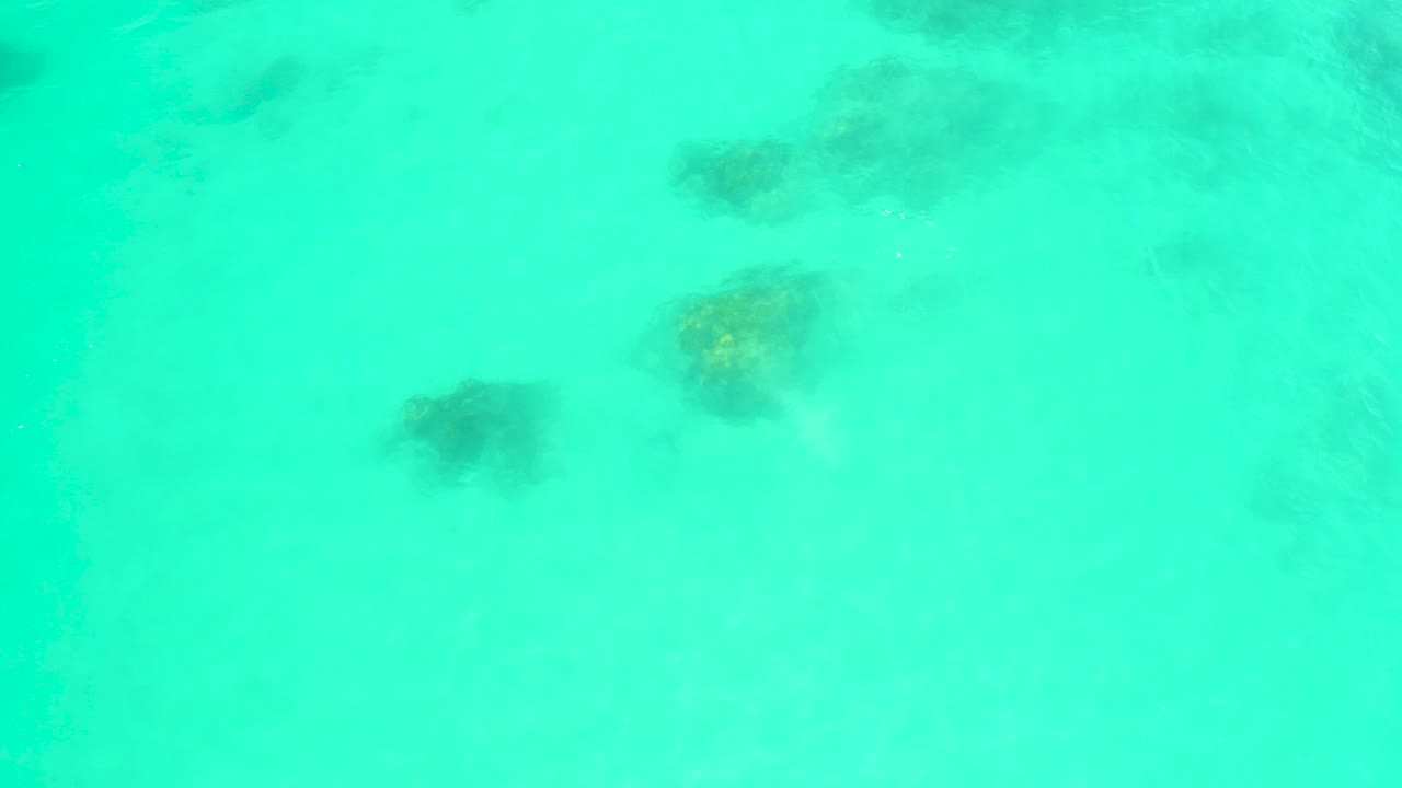 Clear turquoise water at Marquis Los Cabos covers clustered dark rocks and coral, with surface movement slightly blurring the shapes, no visible people.