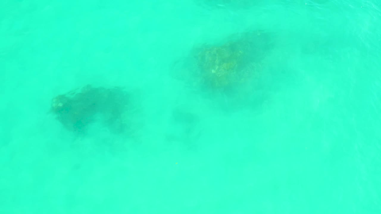 A downward view at Marquis Los Cabos shows scattered dark coral or rock forms beneath hazy turquoise sea water and no visible people.