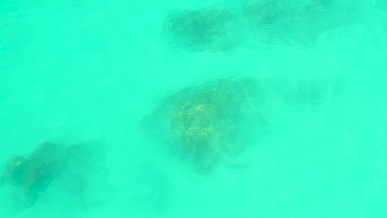 Turquoise water at Marquis Los Cabos covers several dark submerged rocks on a pale seabed, with no visible people.
