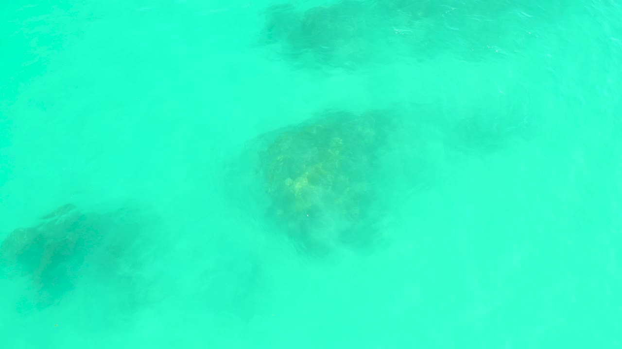 Dark rock patches remain visible beneath shallow turquoise water at Marquis Los Cabos, with a faint bright speck in the water and no visible people.