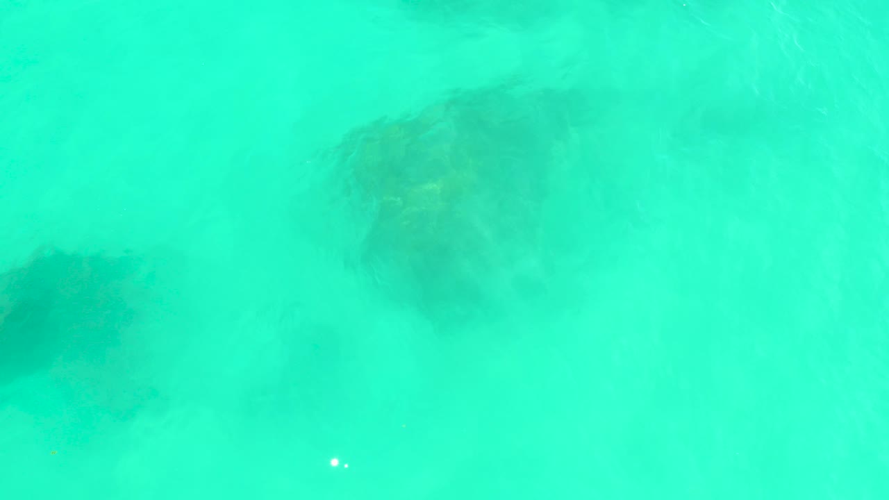 At Marquis Los Cabos in Baja California Sur, turquoise water covers several dark submerged rocks near the top and lower left of the frame, with no visible people.
