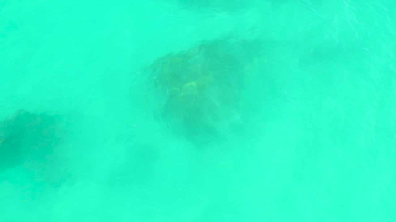 Turquoise water at Marquis Los Cabos reveals a larger dark rock form in the lower left and smaller submerged shapes under soft ripples, with no visible people.