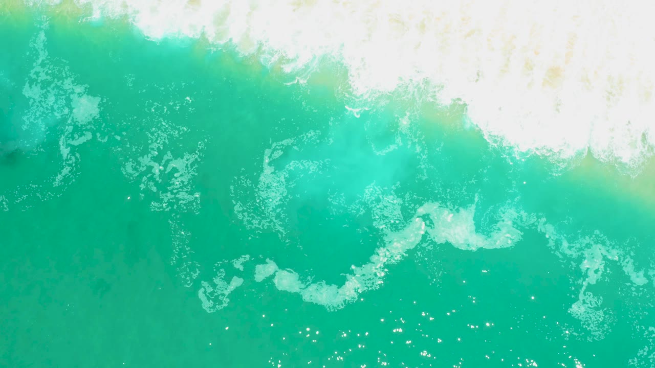 Overhead frame from Marquis Los Cabos with white foam spreading diagonally through turquoise water and small bubbles below, with no visible people.