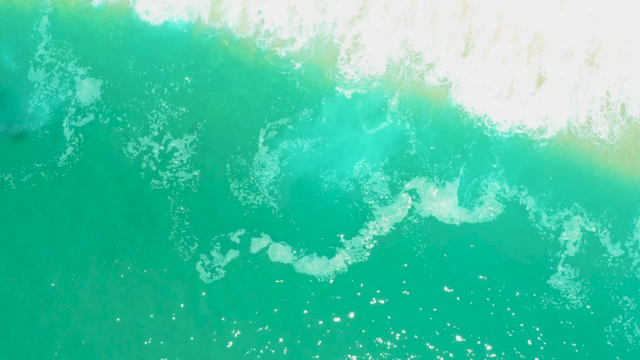 Aerial view at Marquis Los Cabos showing a broken white foam band across turquoise ocean water, with glittering highlights and no visible people.