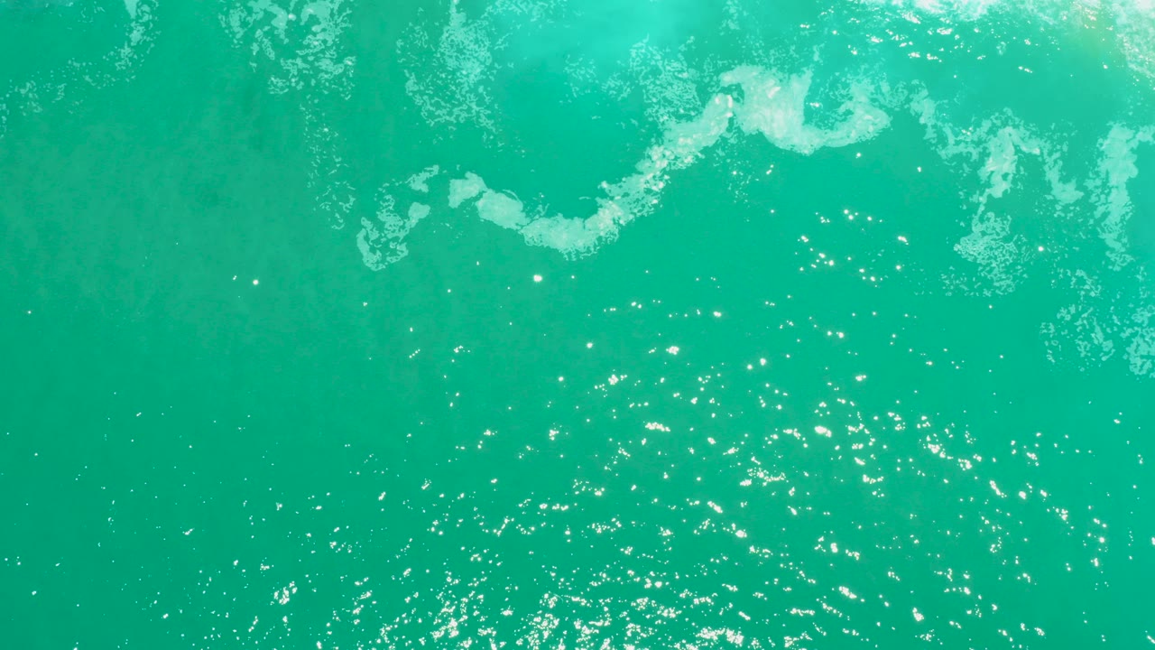 Top-down aerial view of turquoise water at Marquis Los Cabos with scattered white foam and sun glints across the surface, no visible people.