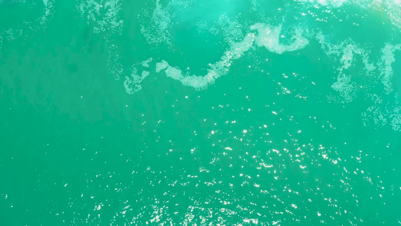 Aerial frame over turquoise ocean water at Marquis Los Cabos, with irregular foam patches near the top edge and bright reflection speckles below, no visible people.