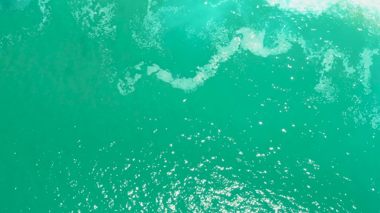 Top-down view of turquoise sea at Marquis Los Cabos with a band of broken white foam along the upper edge and sparkling water below, no visible people.