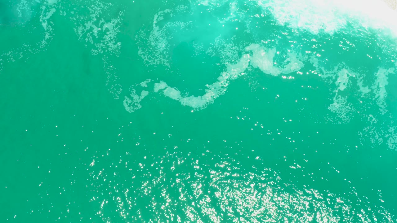 Aerial view at Marquis Los Cabos showing turquoise water, curved white foam patterns, and bright surface reflections with the sandy shore still outside the frame, no visible people.