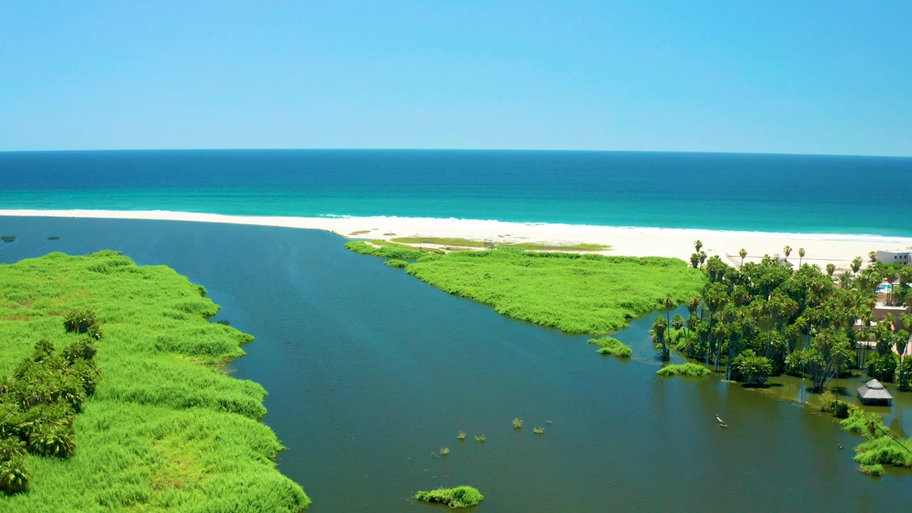 Opening aerial frame over San Jose del Cabo Estuary with dark lagoon water in the foreground, bright green vegetation on both sides, white sand beach, blue ocean beyond, and no visible people.