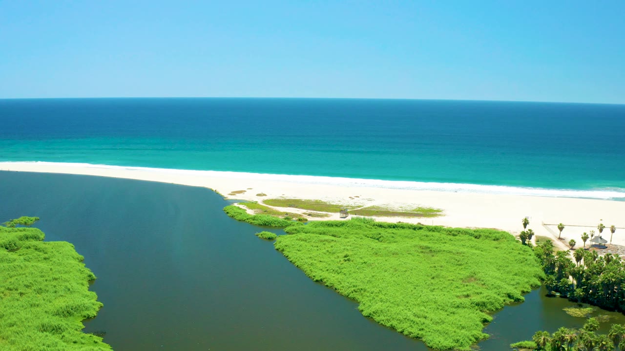 Frame at 0:03 shows San Jose Del Cabo Estuary water along the left, a broad green vegetated bank in the lower frame, white beach across the middle, blue ocean beyond, and no visible people.
