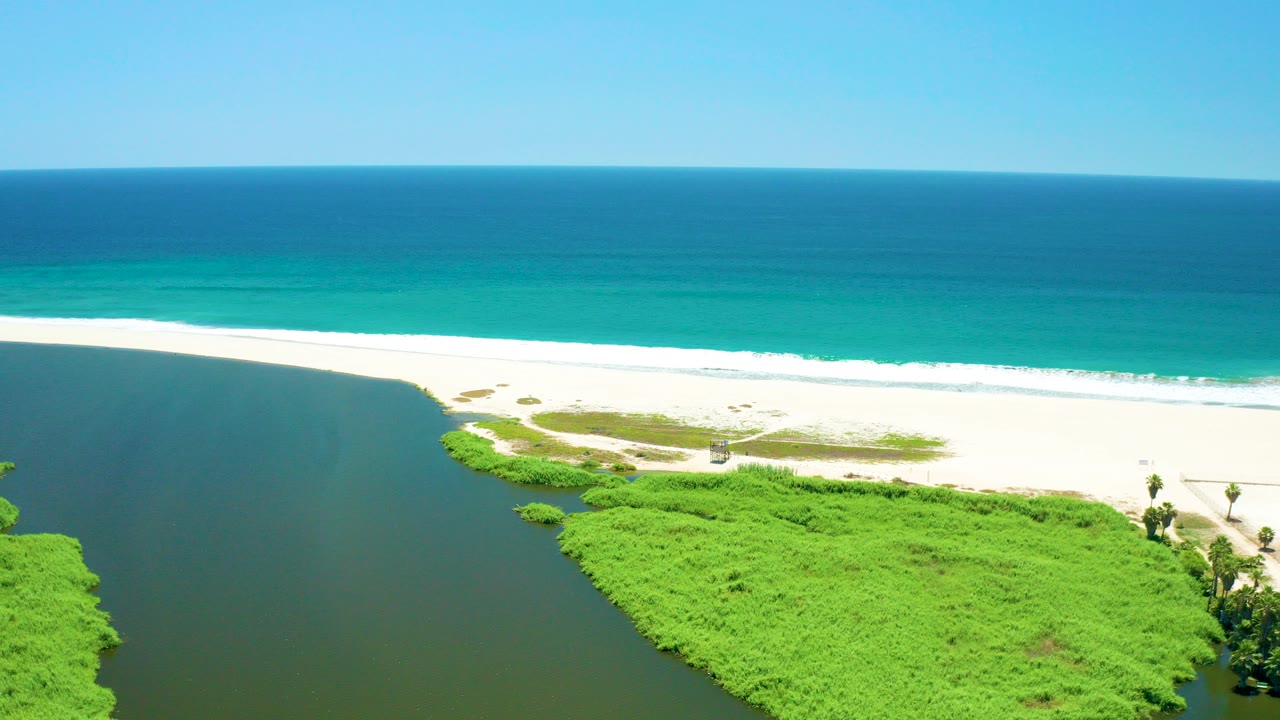Opening aerial frame over San Jose del Cabo Estuary, Mexico, showing dark river water beside bright green vegetation, a white sand beach, turquoise ocean, and no visible people.