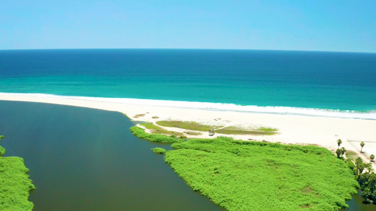 Aerial frame over San Jose del Cabo Estuary, Mexico, with the dark lagoon edge on the left, green vegetation below, white beach through the center, turquoise ocean beyond, and no visible people.