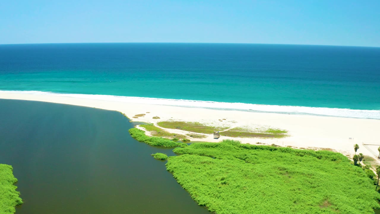 Mid-clip aerial frame at San Jose del Cabo Estuary, Mexico, with dark river water entering from the left, low green vegetation below, pale sand at center, turquoise ocean behind it, and no visible people.