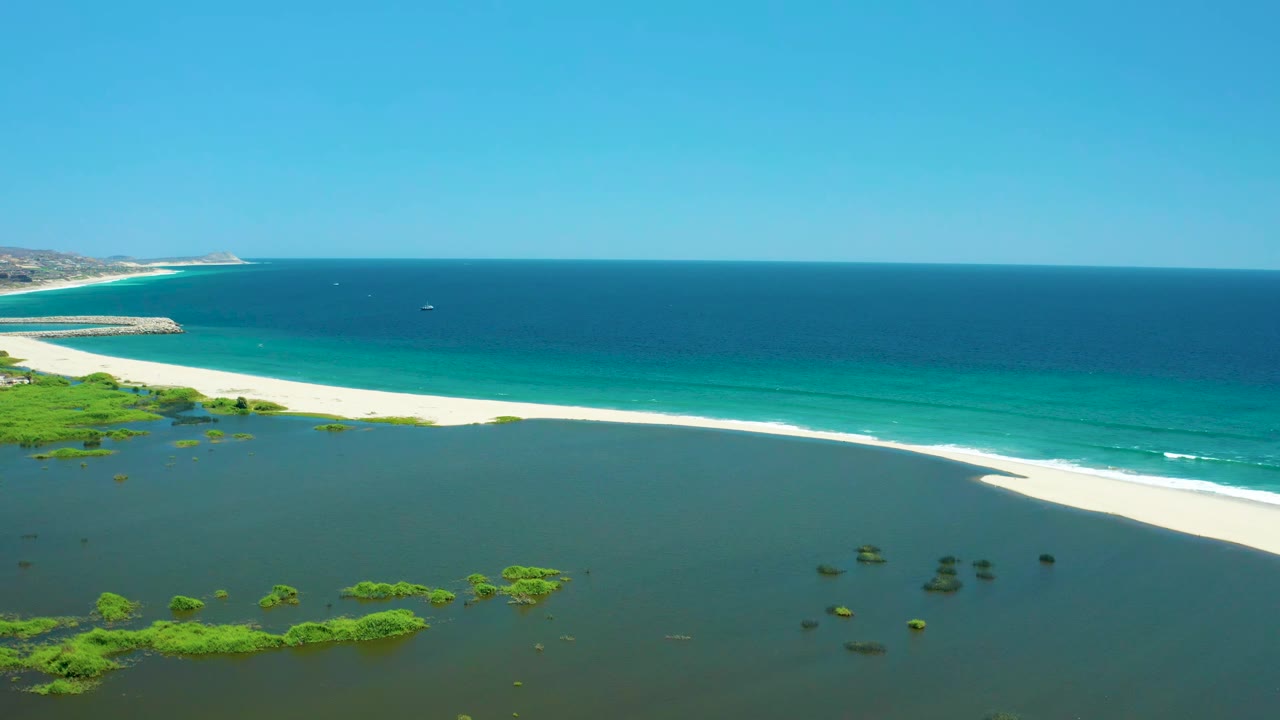 Wide aerial coastline with lagoon plants, white beach, turquoise waves, and open sea.