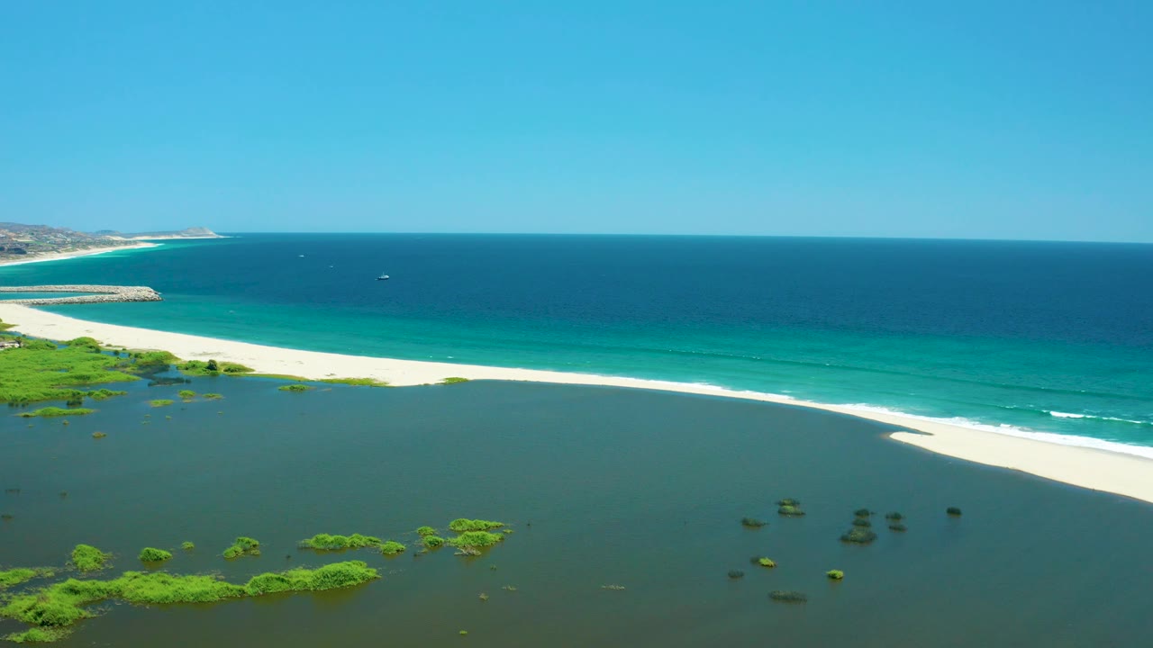Aerial view of San Jose Del Cabo Estuary with scattered green vegetation in shallow lagoon water, a curving white sand bar, turquoise shoreline water, open ocean horizon, and no visible people.