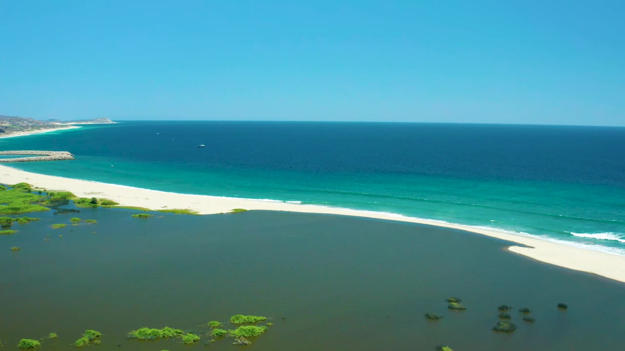 At 0:01, the San Jose del Cabo Estuary shoreline runs diagonally across the frame with green lagoon water below, white sand, blue ocean, a tiny distant boat, and no visible people.