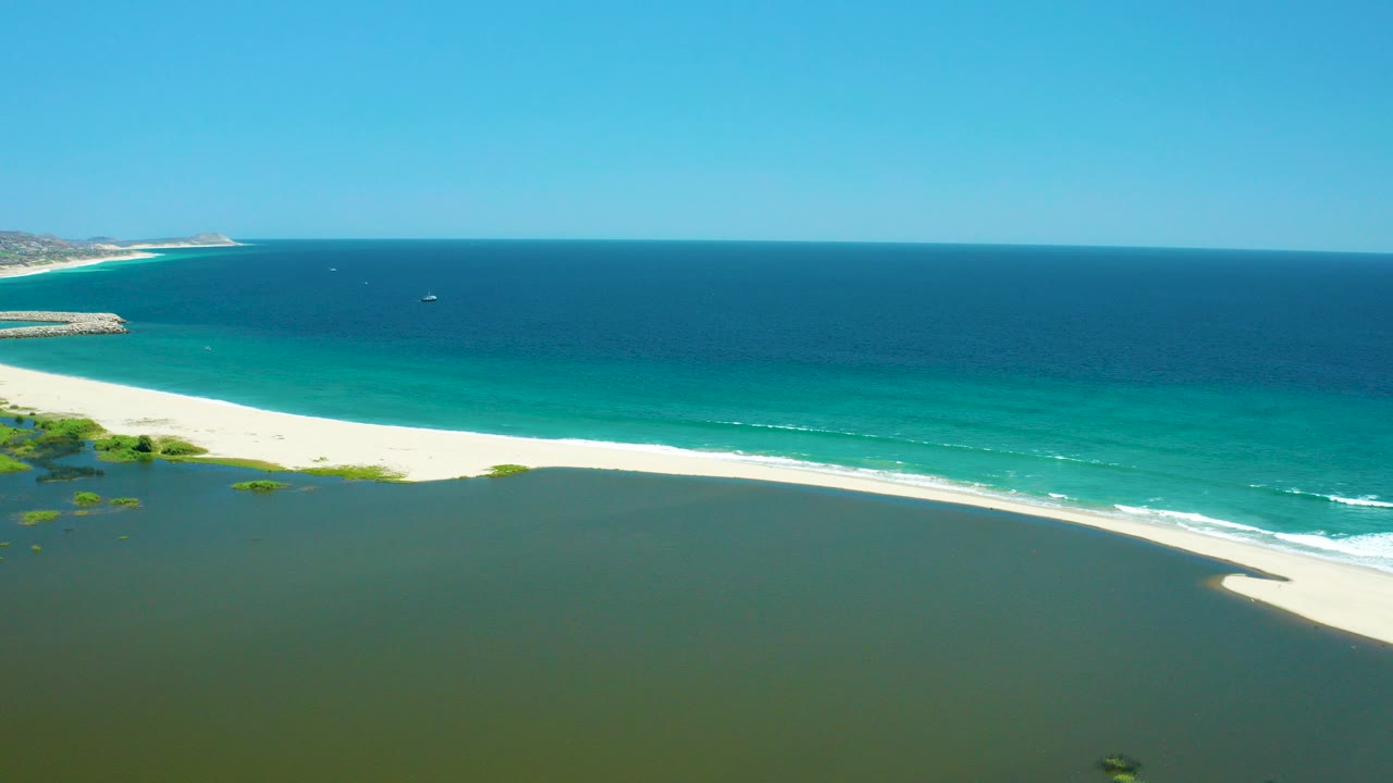 Aerial frame of San Jose del Cabo Estuary with green lagoon water in the foreground, a pale sand beach, turquoise ocean surf, deep blue water beyond, and no visible people.