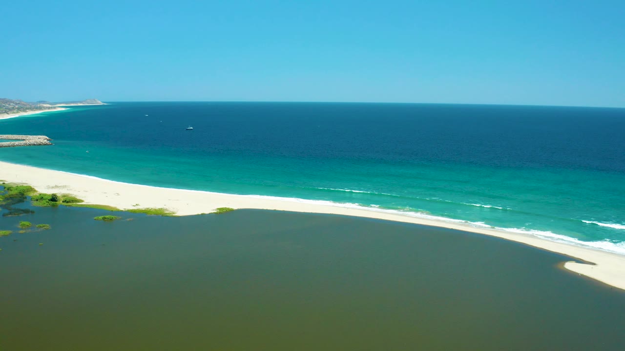 Aerial view of estuary lagoon, white beach, and ocean surf under clear sky.