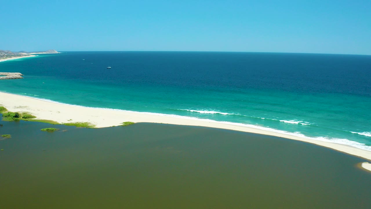 Aerial frame of the San Jose del Cabo Estuary with green water in the foreground, a bright sand bar, turquoise surf, deep blue ocean, distant boats, and no visible people.
