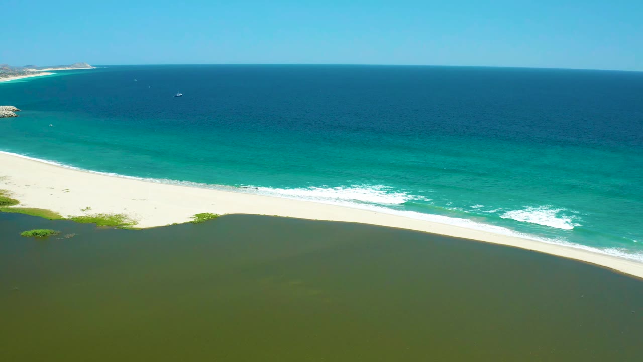 Aerial frame at San Jose Del Cabo Estuary with white foam spreading along the sandy shore, green lagoon water below, blue ocean beyond, small distant boats, and no visible people.