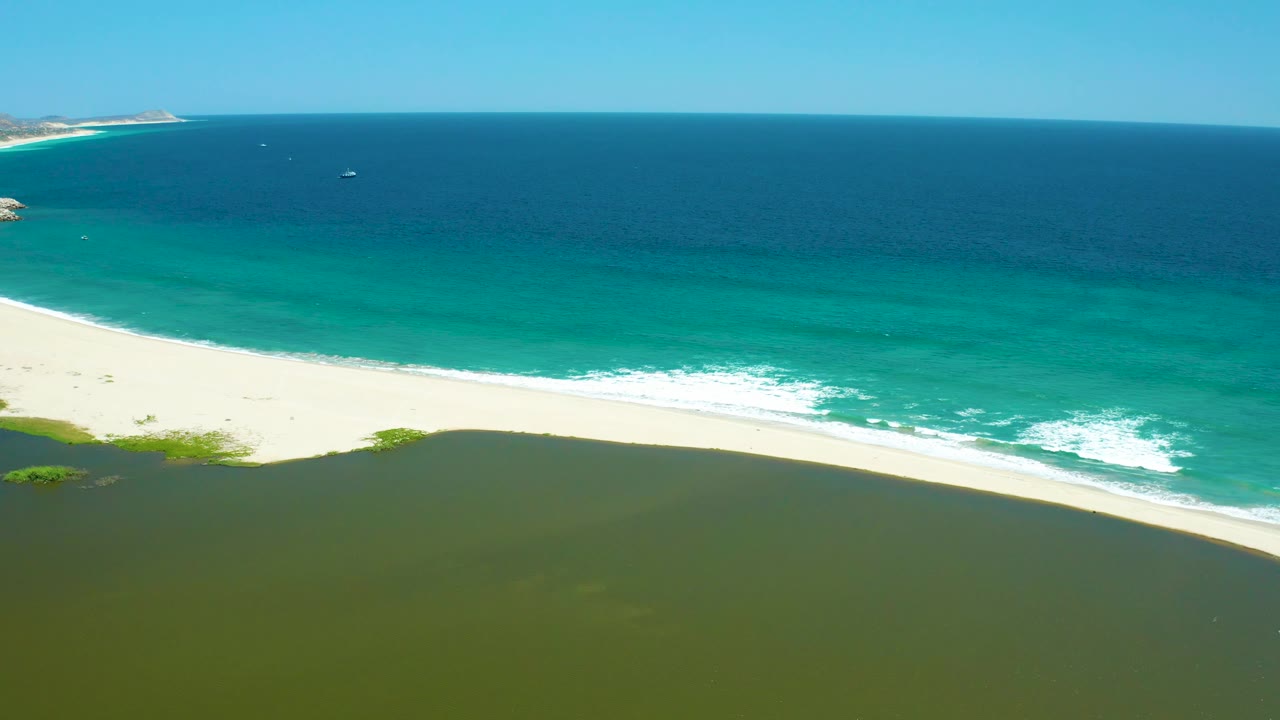 Aerial coastal frame from San Jose Del Cabo Estuary with lagoon water filling the lower frame, waves rolling onto the pale sand, open ocean behind, distant boats, and no visible people.