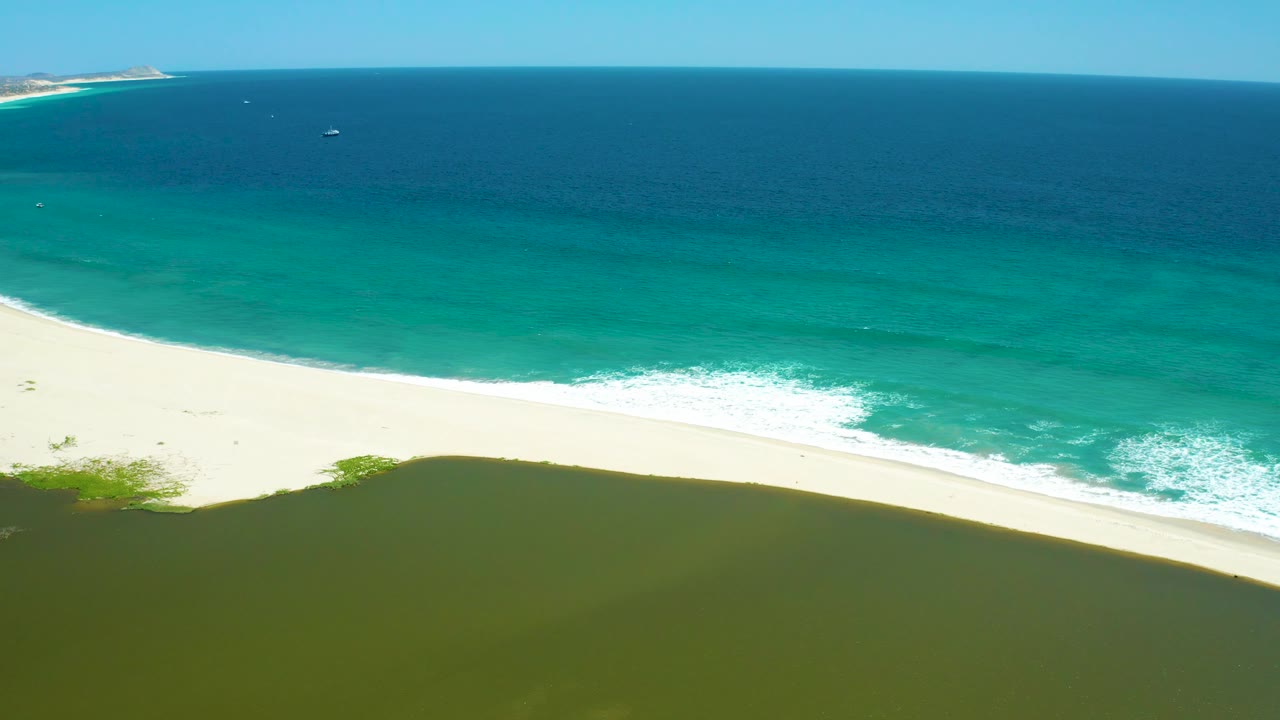 Aerial view over San Jose Del Cabo Estuary with a wide strip of white sand, foamy turquoise surf, dark lagoon water in the lower frame, and no visible people.