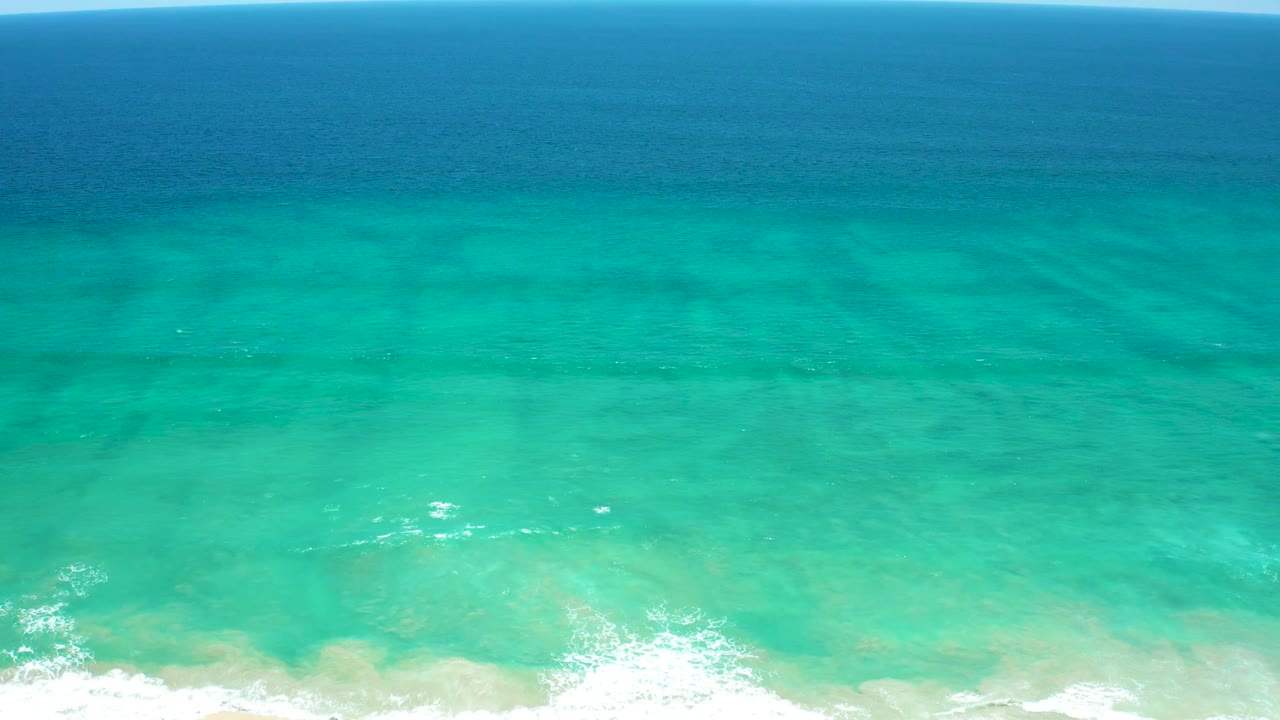 Aerial frame over San Jose Del Cabo Estuary coastal water, with pale sandy shallows under turquoise water and white wave foam thinning across the bottom edge, no visible people.