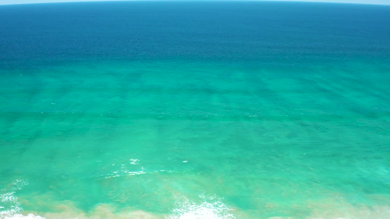 San Jose Del Cabo Estuary ocean surface seen from above, with smooth teal shallows, faint white foam near the lower edge, and a darker blue band toward the horizon, no visible people.