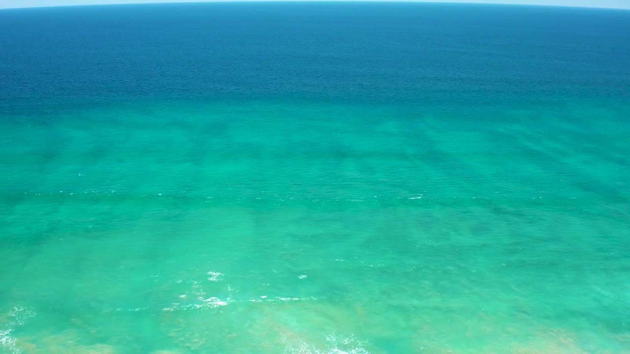 Aerial view at San Jose Del Cabo Estuary with deep blue water across the upper frame, turquoise shallows below, and a thin bright strip of wave foam near the shoreline, no visible people.