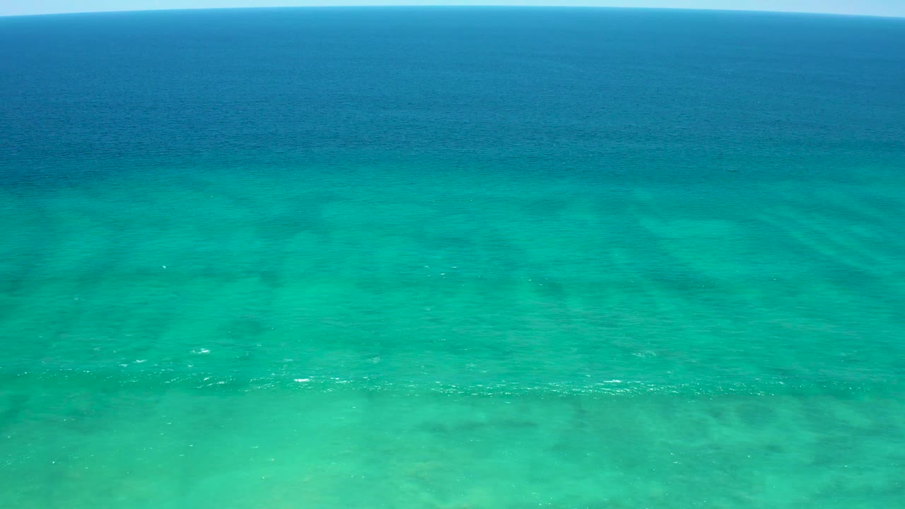 Aerial frame of clear turquoise water in Baja California Sur, with faint white foam streaks across the lower water and darker blue sea along the top, no visible people.