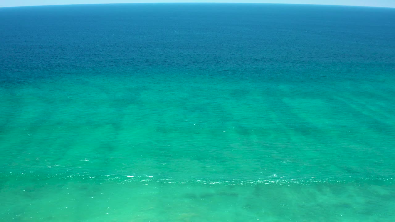 Wide drone view of shallow turquoise ocean water near San Jose del Cabo Estuary, with pale seabed patterns, scattered foam flecks, and no visible people or boats.