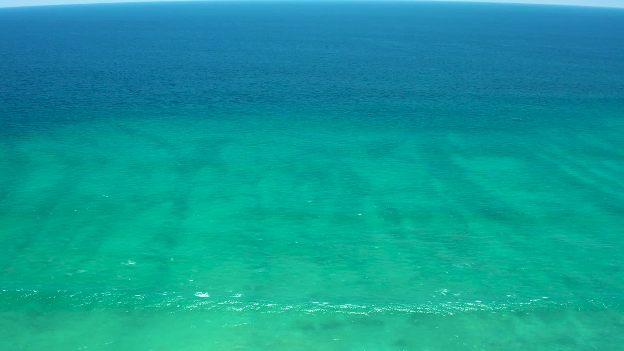 Aerial ocean frame from San Jose del Cabo Estuary showing turquoise foreground water, a horizontal darker blue band near the horizon, subtle ripples, and no visible people.