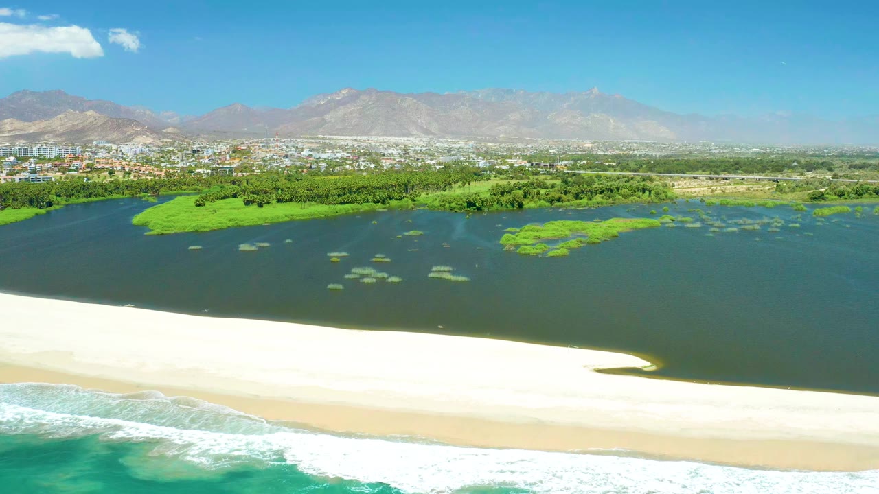 Aerial view over San Jose Del Cabo Estuary with foamy ocean in the foreground, a bright sandy beach, dark lagoon water, green vegetation, distant buildings, hazy mountains, and no visible people.