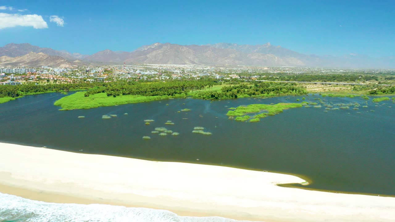 Aerial view of sandy shore, lagoon water, green vegetation, city, and mountains.