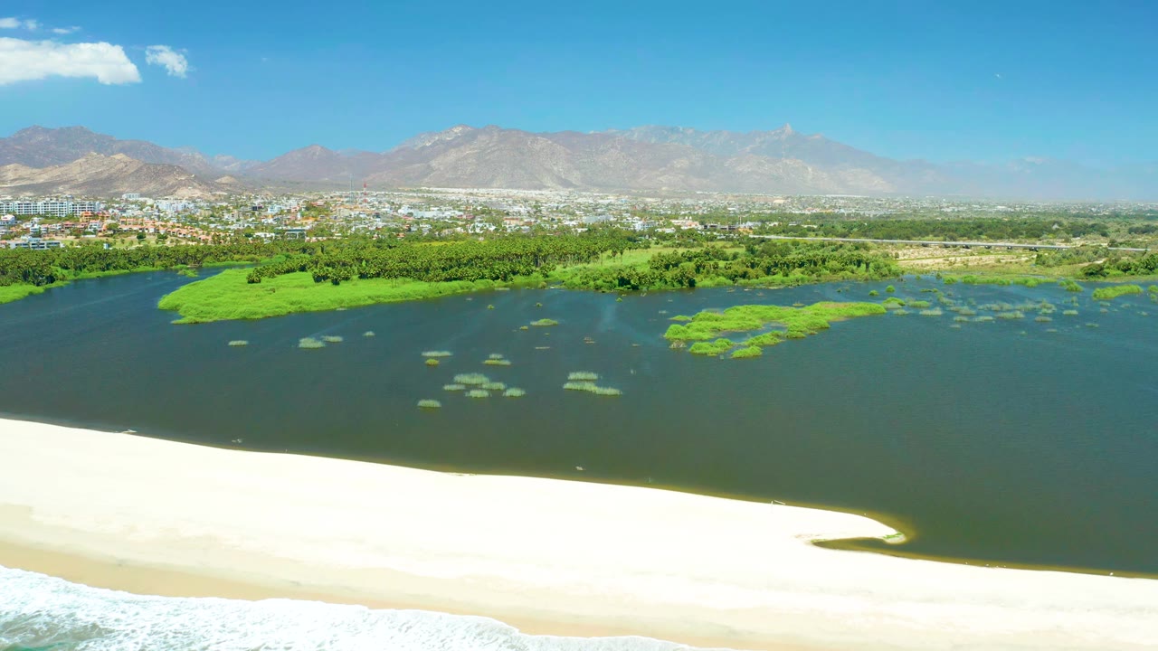 At 0:01, the San Jose Del Cabo Estuary lagoon fills the frame behind a bright sandbar, with green floating islands, a city strip, hazy mountains, clear sky, and no visible people.