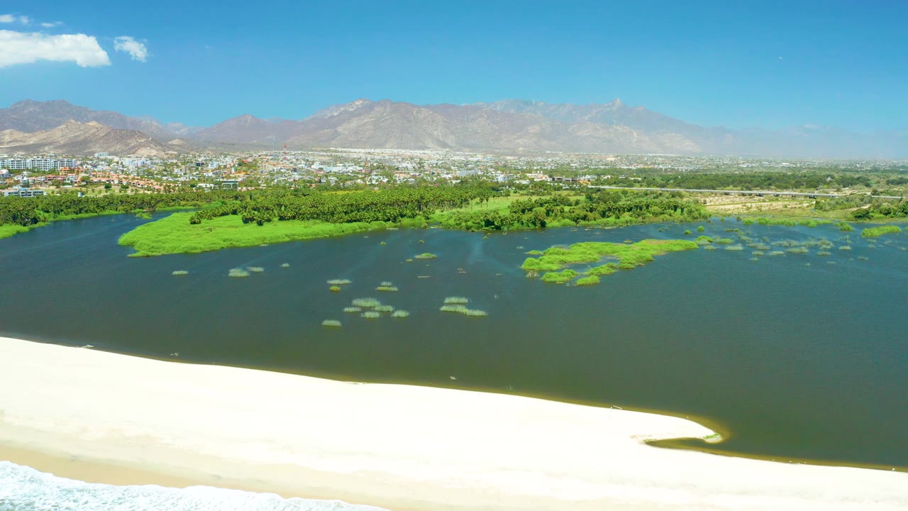 At 0:02, the aerial view holds on the San Jose Del Cabo Estuary with dark water meeting pale sand, scattered green vegetation in the lagoon, distant city blocks, mountains, and no visible people.