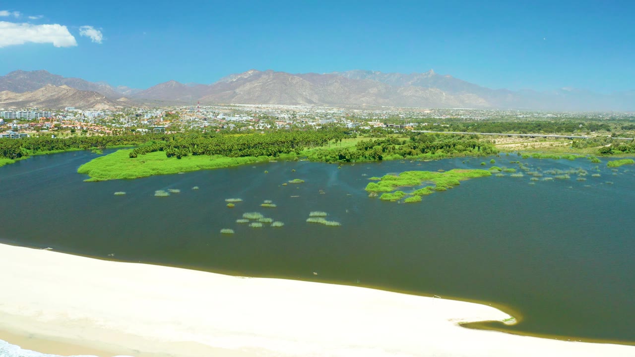 At 0:03, San Jose Del Cabo Estuary appears from above with the sandy shore low in frame, broad lagoon water, bright green patches, a city band, hazy mountains, blue sky, and no visible people.