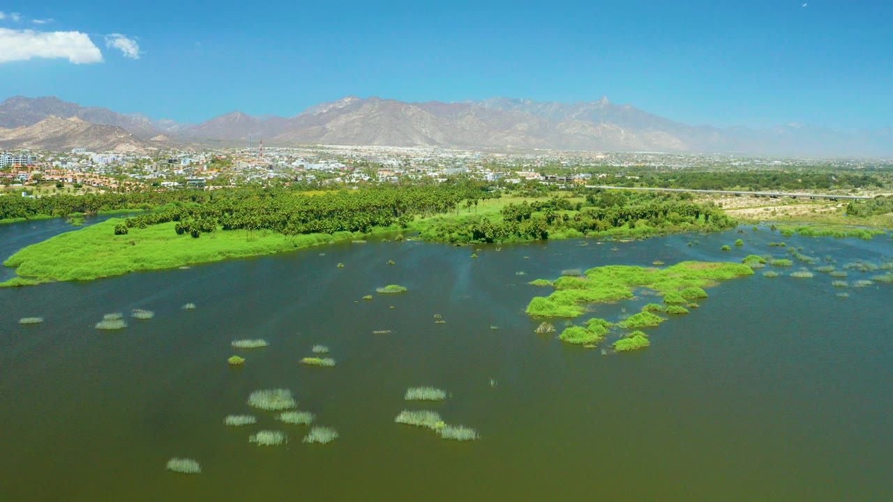 At 0:00, an aerial view over San Jose Del Cabo Estuary shows dark water with scattered green vegetation, a tree belt, low buildings, distant mountains, blue sky, and no visible people.
