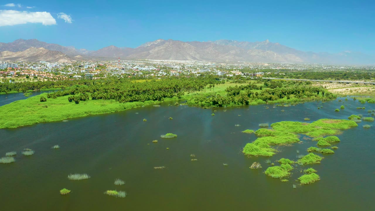 San Jose Del Cabo Estuary aerial with more open water centered in frame, bright green mats on the right, a low city band behind trees, hazy mountains, blue sky, and no visible people.